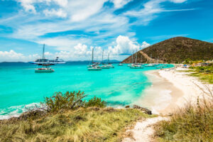 Turquoise Caribbean bay with anchored sailboats and a cruise ship, pristine white sand beach, and lush green hillside under blue sky.