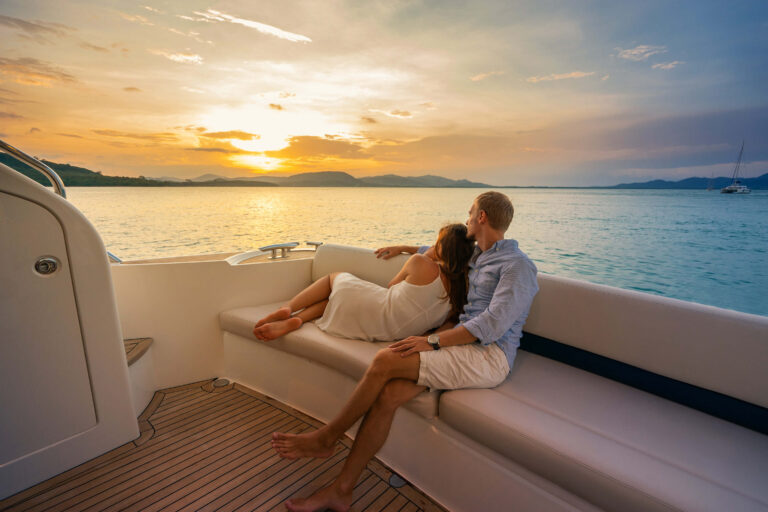 Couple relaxing on luxury yacht deck at sunset in tropical waters with island mountains in background.