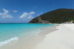 A pristine white sand beach with turquoise waters meets a lush green hillside under a blue sky with white clouds.