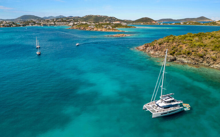 Catamaran anchored in turquoise waters off a Mediterranean coastal town with hills and sailboats in the background.