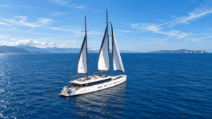 Luxury gulet sailing yacht with white sails in Mediterranean waters near mountainous Greek islands under blue sky.