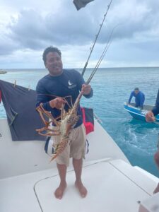 Man holding large lobster on fishing boat with turquoise ocean water and blue sky in background.