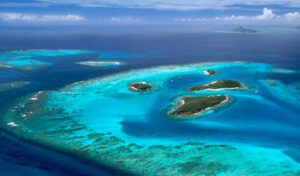 Aerial view of turquoise tropical waters with small islands and a yacht anchored in a pristine atoll lagoon.