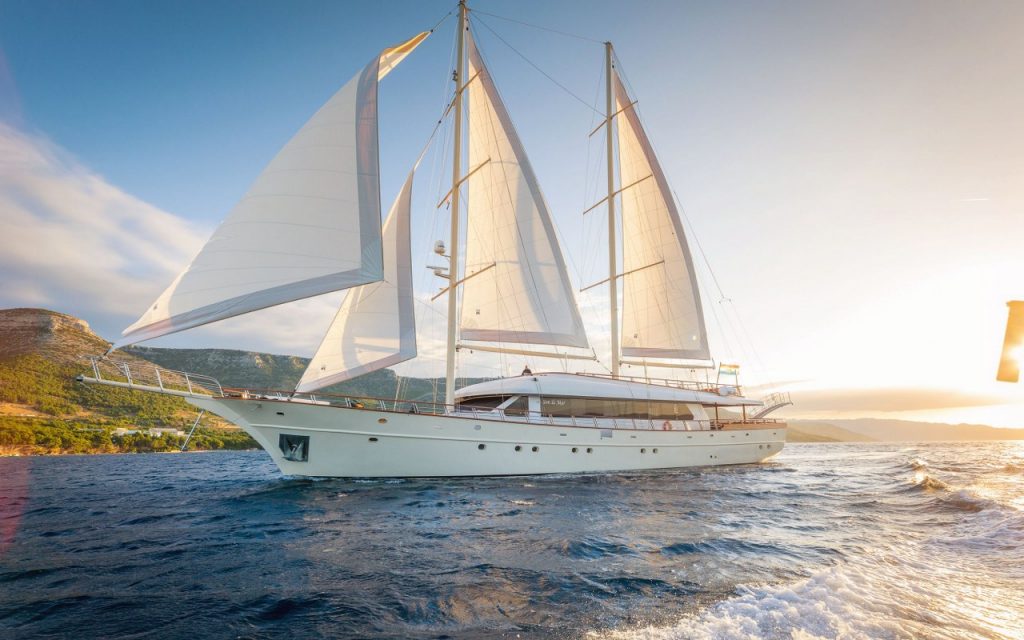 Luxury gulet sailing yacht with white sails anchored in Mediterranean bay at sunset with mountainous coastline.